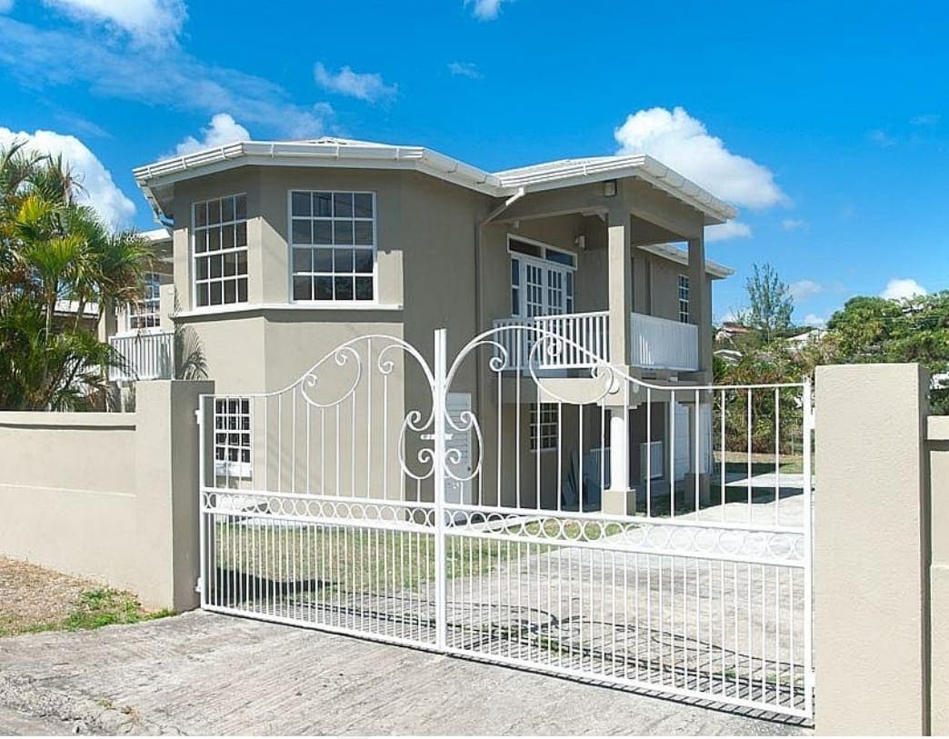 A modern two-story apartment building is seen behind a decorative white metal gate. Large windows enhance the building's facade, and the driveway is visible, providing access to the covered parking area beneath the elevated structure.