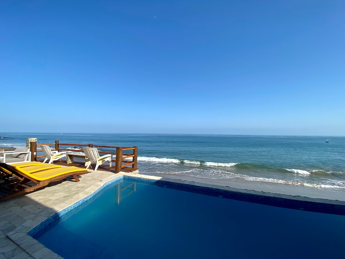 A clear view of the ocean is presented, with gentle waves lapping at the shore. A pool reflects the sky, and lounge chairs are positioned on the deck, inviting relaxation. The horizon shows a seamless blend of sea and sky under bright daylight.