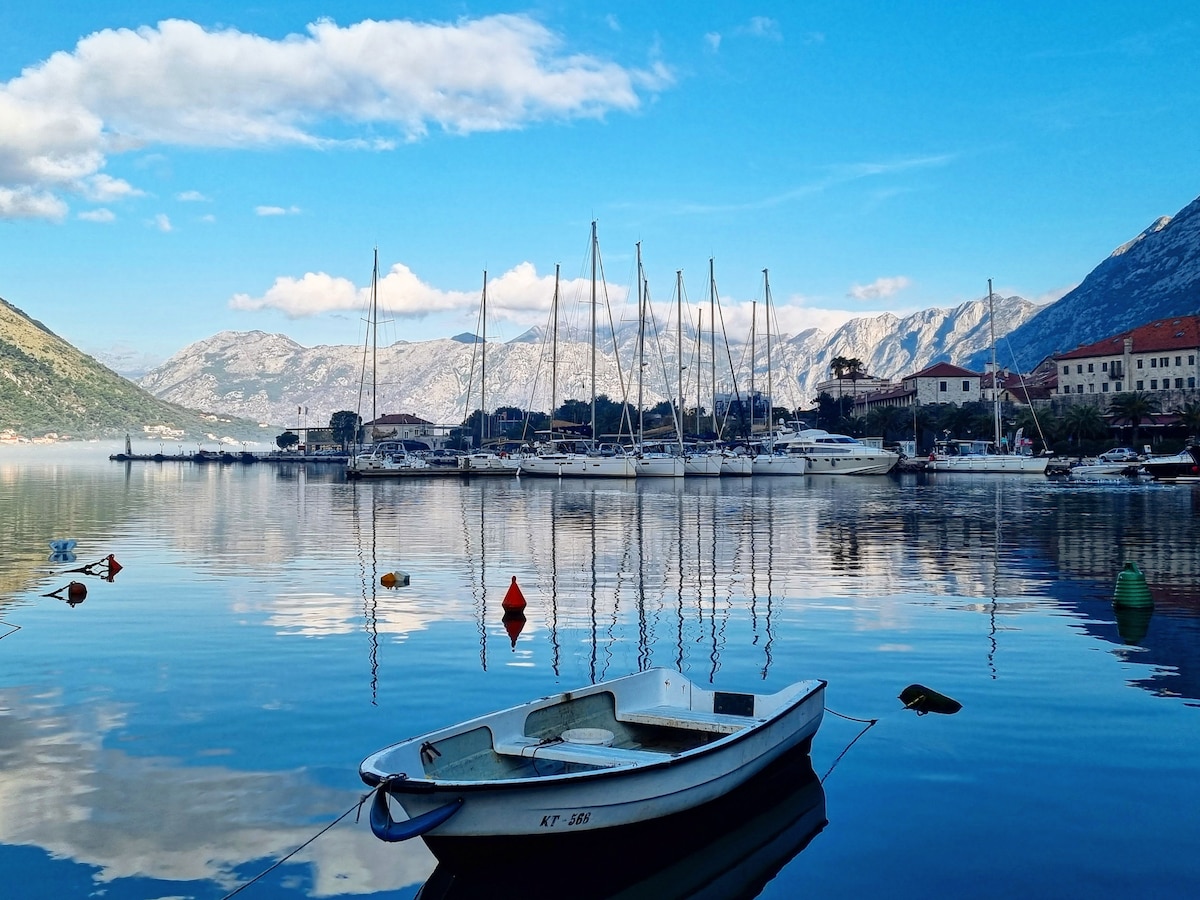 A serene view of a calm bay is presented, featuring a small boat floating gently on the water. Sailboats are docked nearby, with mountains framing the scene, while reflections of the boats and coastal structures can be seen on the surface.
