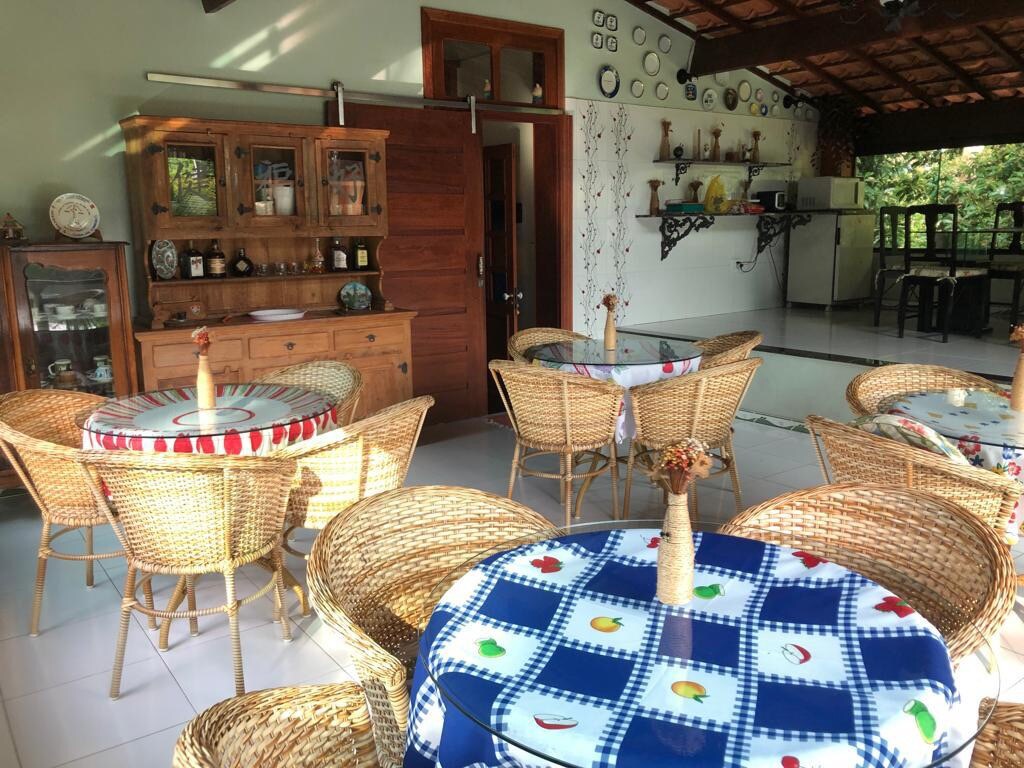 The dining area features several round woven chairs surrounding tables covered with colorful tablecloths. A wooden cabinet is positioned against the wall, showcasing decorative items. Natural light fills the space, enhancing the open layout leading to a kitchen area in the background.