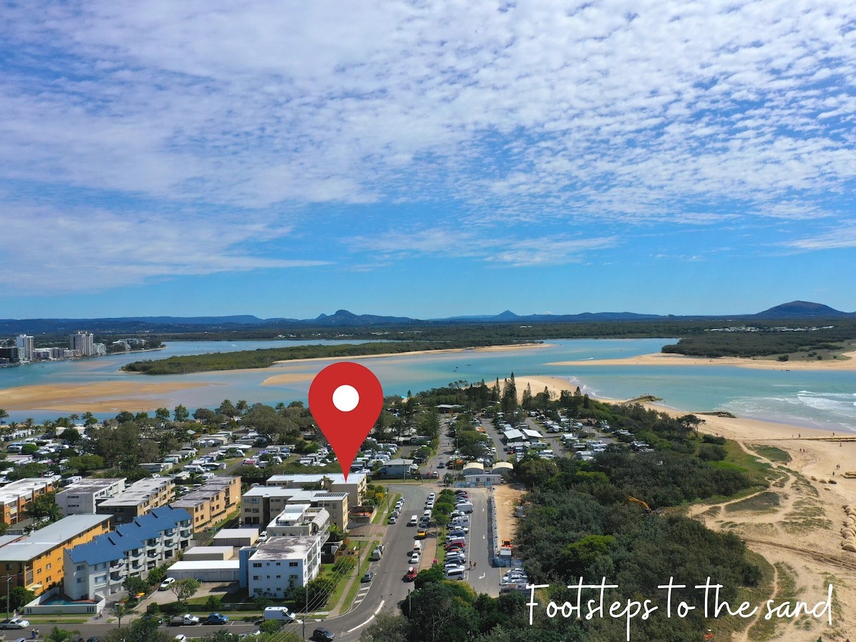 An aerial view showcases the beachfront location of 'Footsteps to the Sand,' surrounded by lush greenery and sandy shores. The nearby Maroochydore River is visible, with its calm waters contrasting the ocean. The vibrant landscape is dotted with residential buildings and open spaces, indicating a welcoming area.