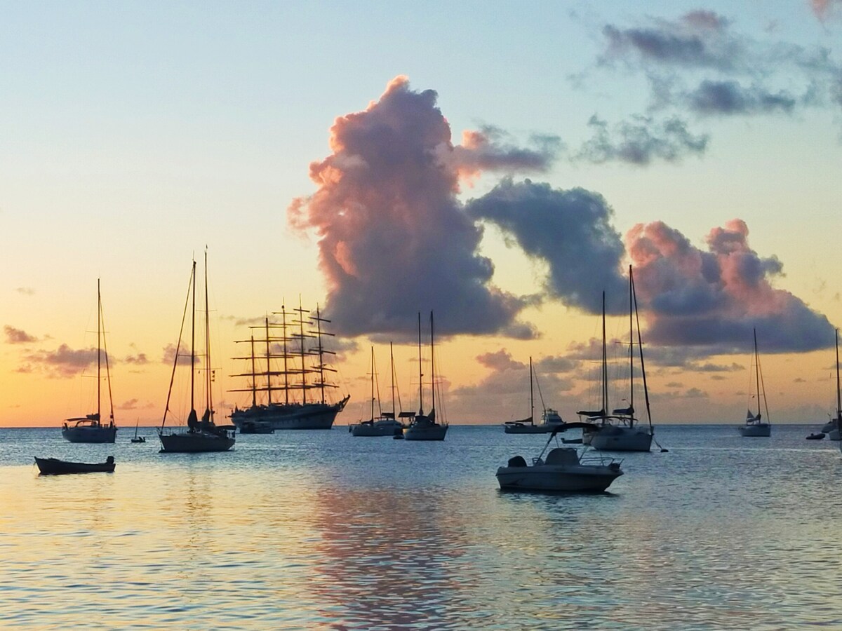 A serene seascape captures various boats anchored in calm waters at sunset. A tall ship with multiple sails is prominently featured, accompanied by smaller vessels. Soft hues of orange, pink, and purple in the sky reflect on the water's surface, creating a tranquil atmosphere.