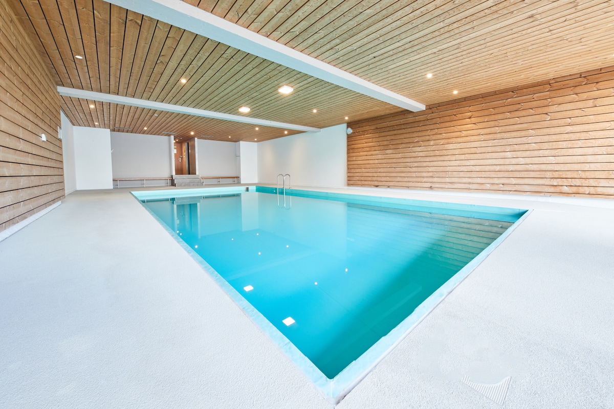 An indoor swimming pool is framed by wooden paneling and illuminated by overhead lights. The tranquil water is calm and inviting, with steps leading into the pool at one end. The surroundings feature a clean, minimalist design with a spacious feel.