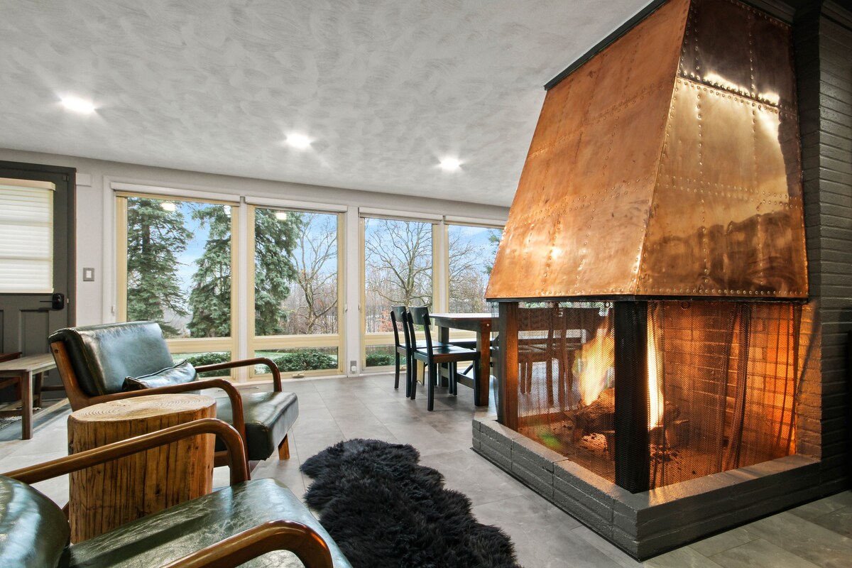 An inviting living area is highlighted by a unique copper fireplace, providing warmth and a focal point. Large windows invite natural light, offering views of the surrounding greenery. Comfortable seating options are arranged for relaxation, complemented by a stylish dining table nearby.