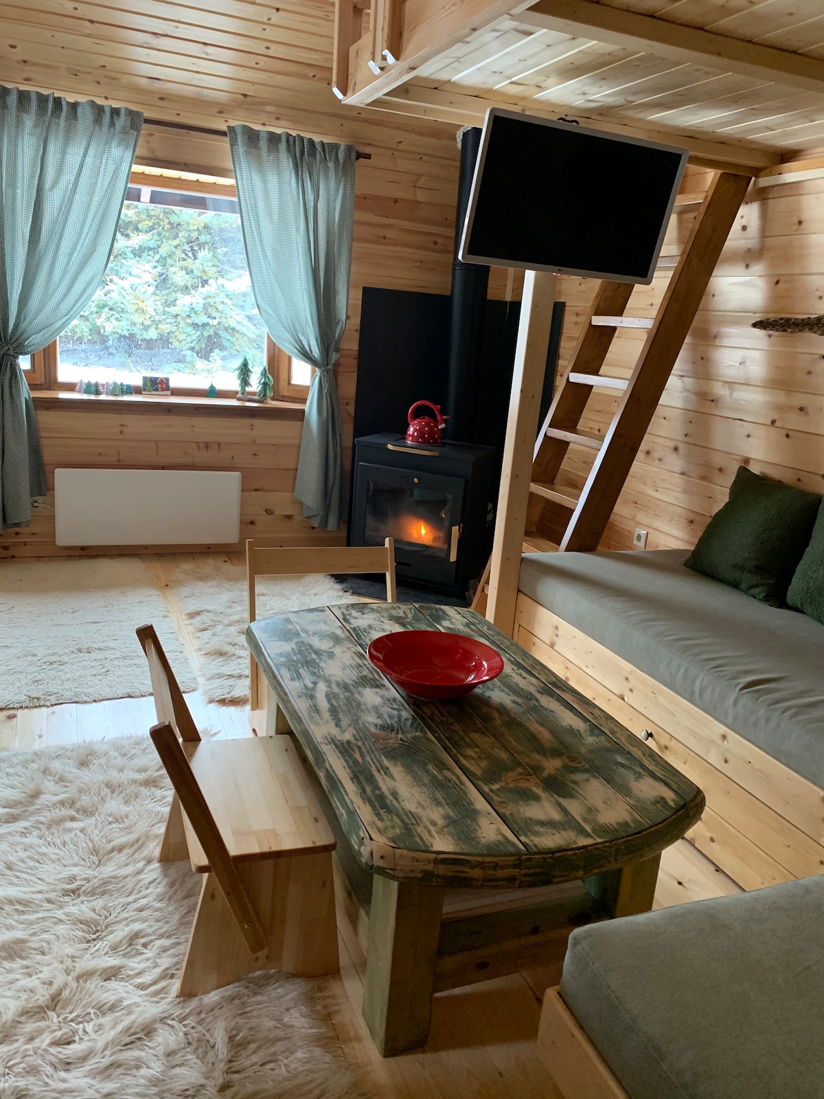 A wooden interior features a cozy sitting area with a compact table and two chairs. A flat-screen television is mounted on the wall. Natural light enters through a window adorned with light curtains, and a small electric heater offers additional warmth.