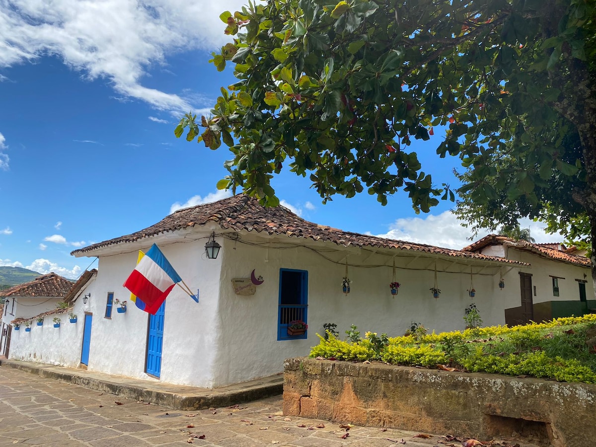 A charming colonial house is visible, featuring whitewashed walls and a terracotta roof. Decorative elements include colorful flowers and a flag displayed at the entrance. Lush greenery surrounds the structure, enhancing the appeal of the neighborhood streets under a clear blue sky.