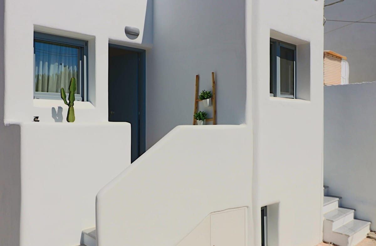 The exterior of the apartment is presented, showcasing smooth white walls typical of Cycladic architecture. A set of stairs leads to the entrance, flanked by a small cactus on a table and a decorative plant on a ladder, contributing to the minimalist aesthetic.