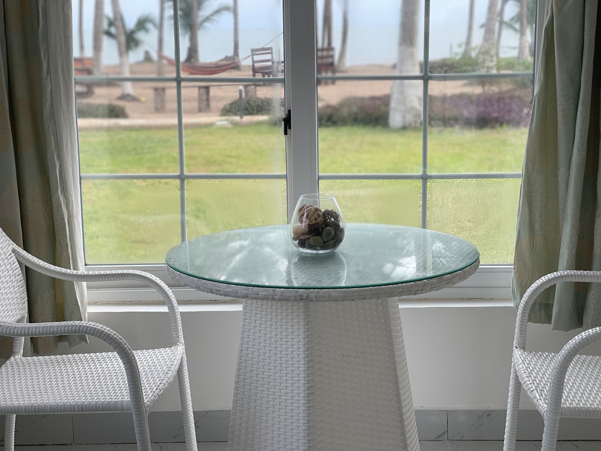 A round glass table is positioned beside a large window, surrounded by two white chairs. The view outside features a lush green lawn and palm trees, with glimpses of the ocean beyond. Natural light gently illuminates the space.
