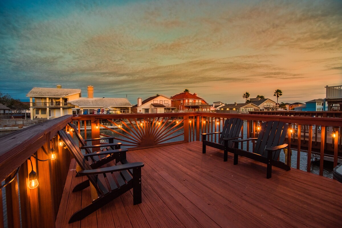 An elevated deck is illuminated by warm string lights, featuring several black wooden chairs arranged for relaxation. The backdrop showcases colorful houses along the canal under a serene sunset sky, enhancing the inviting outdoor space.