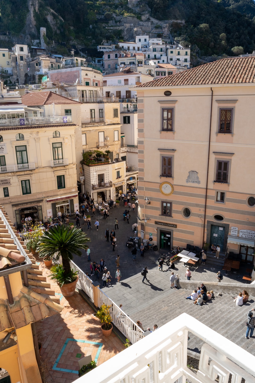 A view of the bustling Piazza Duomo in Amalfi is captured, showcasing vibrant streets lined with buildings of various architectural styles. People can be seen enjoying the open space, while palm trees and outdoor seating areas add to the lively urban atmosphere.