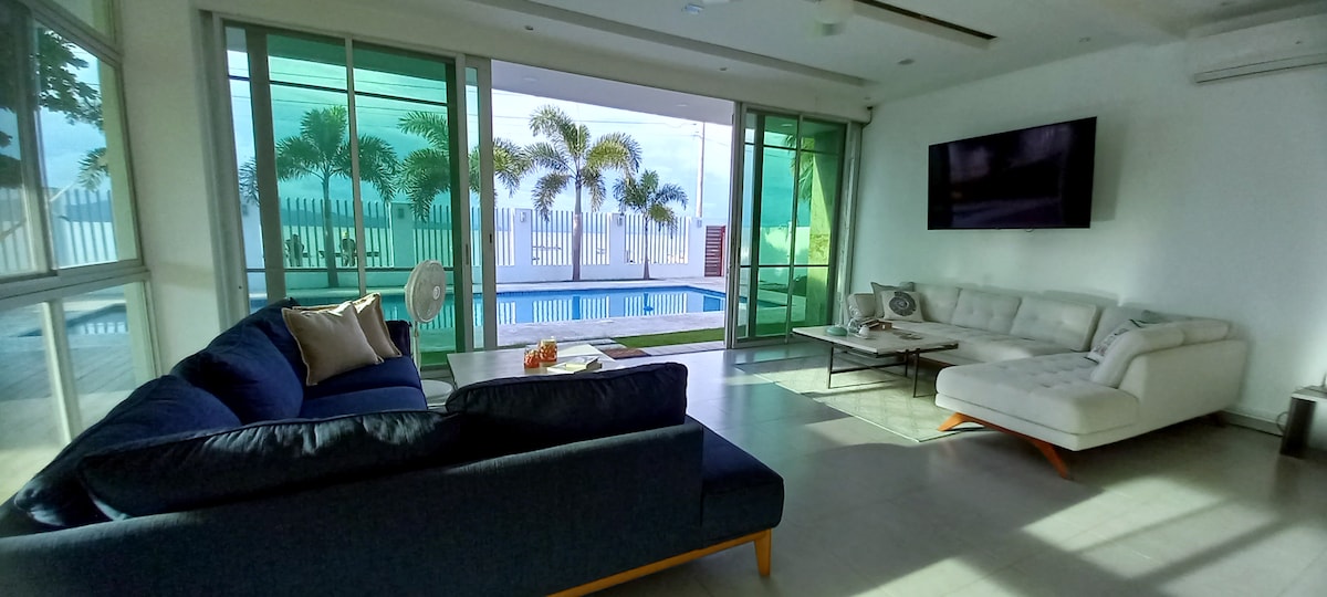An open living area features large glass doors that reveal a view of palm trees and a private pool. A spacious blue sofa and a light-colored sectional couch provide comfortable seating. A wall-mounted TV is positioned above a low coffee table, inviting relaxation.