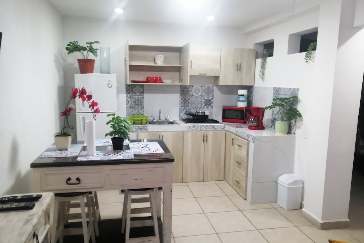 A well-equipped kitchen features wooden cabinets, a white refrigerator, and a microwave. A small table with two stools is positioned in front, adorned with potted plants. Decorative tiles add interest to the backsplash, while under-cabinet lighting enhances the workspace.