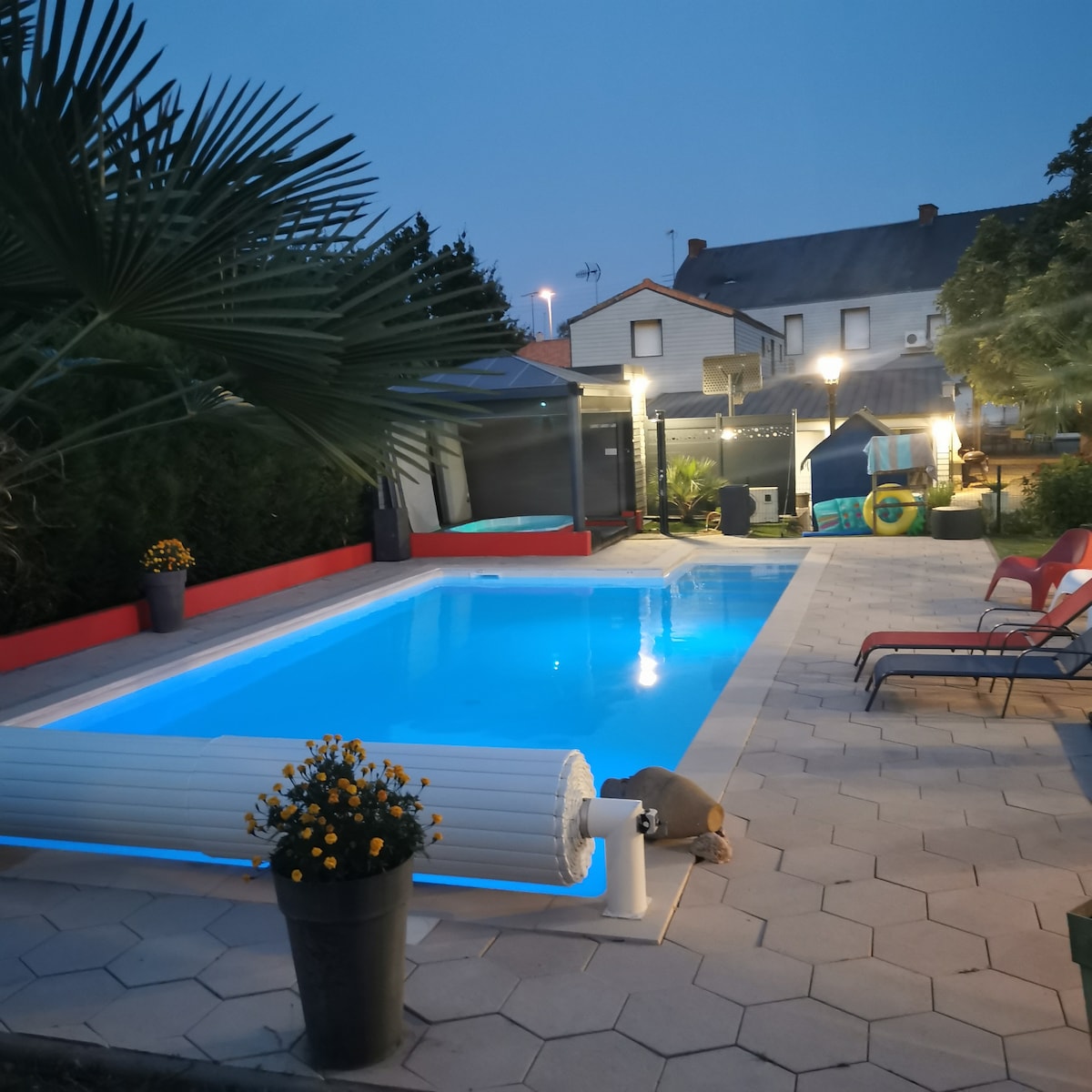 A well-lit swimming pool is surrounded by a tiled patio, showcasing a tranquil blue hue. Sun loungers are positioned nearby, complemented by a planter filled with yellow flowers. Modern lighting enhances the evening ambiance, with greenery providing a natural touch.