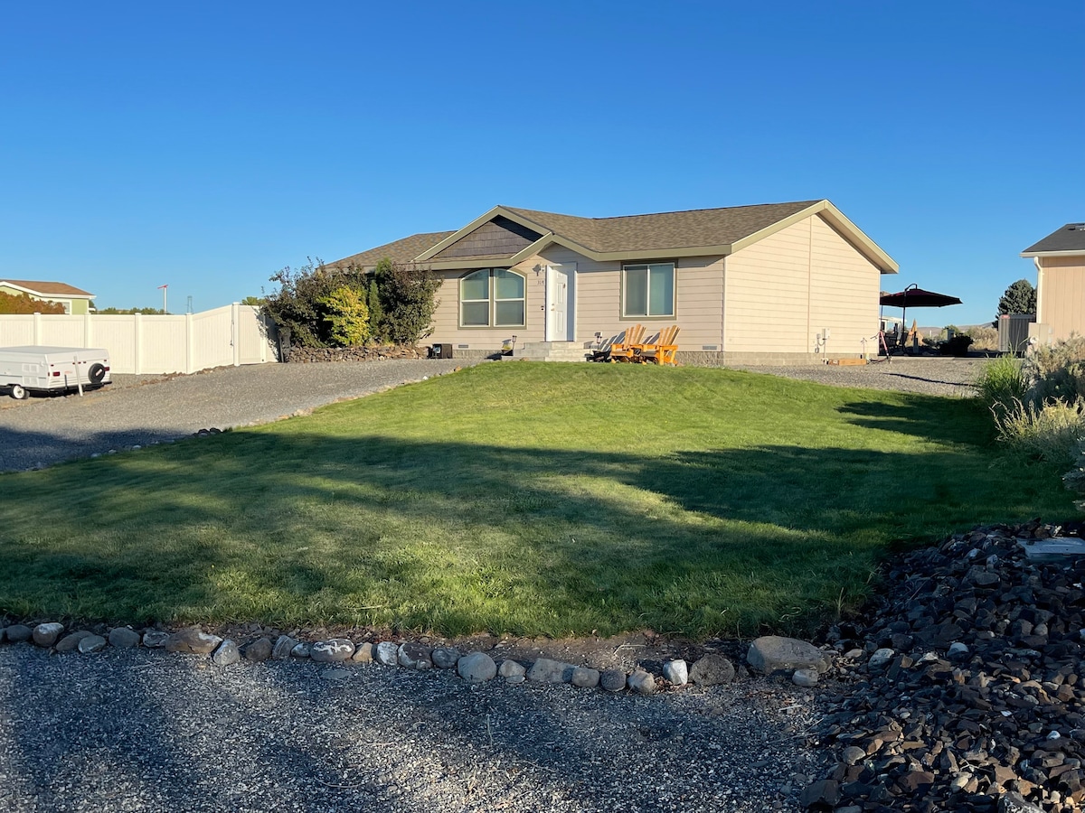 A single-story home with light-colored siding is set against a clear blue sky. A well-maintained lawn provides a green contrast in the foreground, while a gravel driveway leads up to the entrance. Adjoining areas feature shrubbery and outdoor seating options.