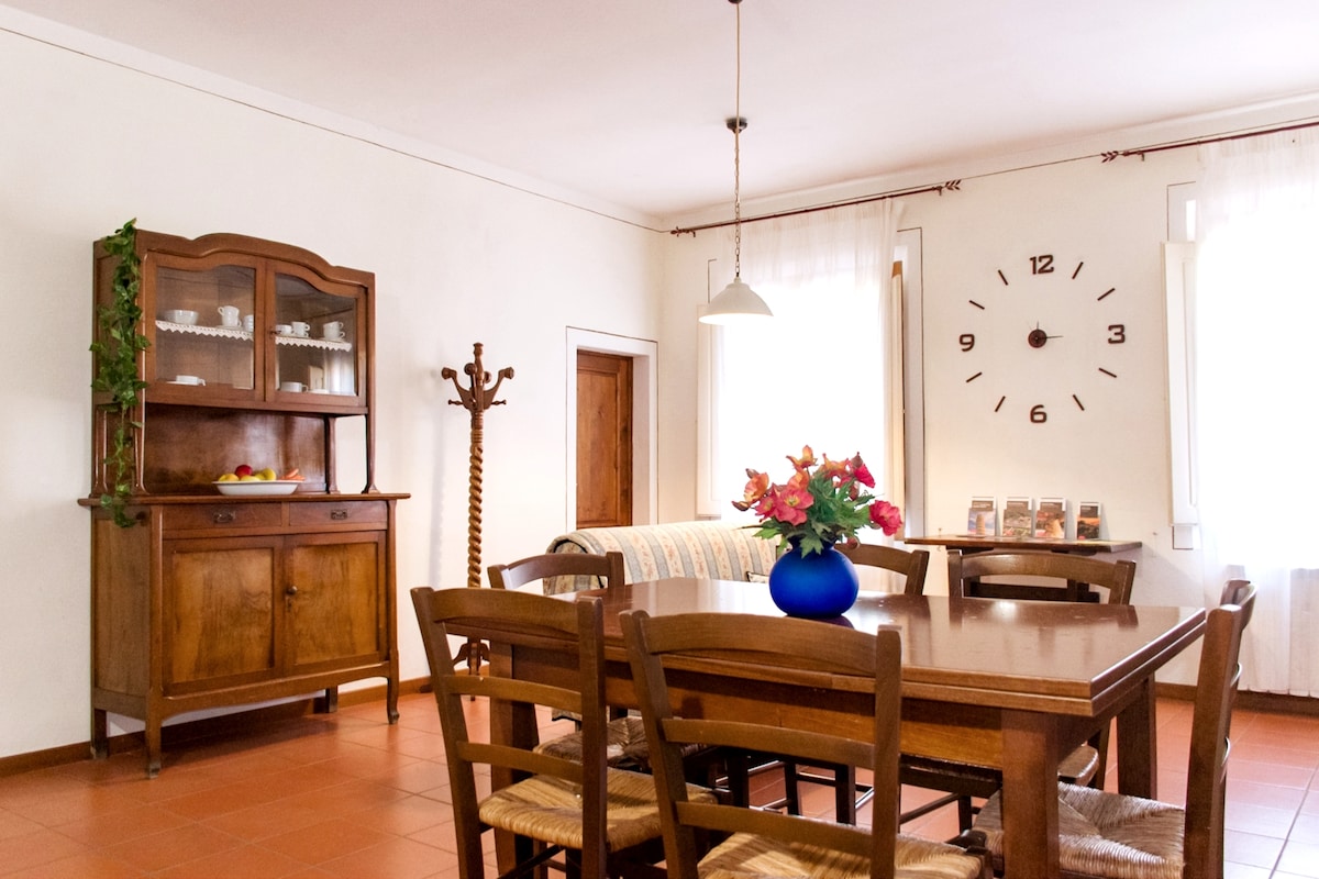 A dining area features a large wooden table surrounded by six chairs, complemented by a vintage cabinet and a blue vase with flowers. Light enters through multiple windows, illuminating a wall clock showing the time. A coat rack stands nearby.