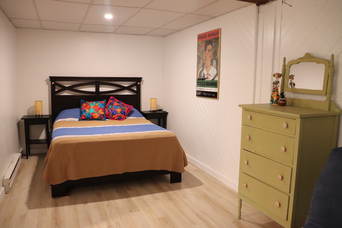 A cozy sleeping area features a spacious bed with a neutral-colored blanket and decorative pillows displaying vibrant patterns. Nightstands with lamps are positioned on either side of the bed. A green dresser adds a pop of color, while an art poster decorates the wall.