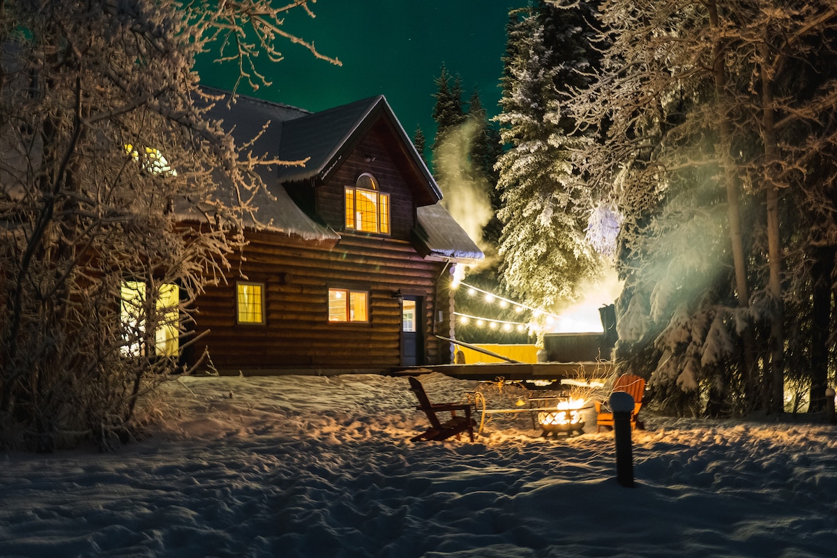 The lodge is illuminated against a snowy backdrop, showcasing warm lights from the windows. A cozy fire pit with seating is visible in the foreground, surrounded by snow-covered trees. The smoke from a nearby jacuzzi rises softly into the night sky.