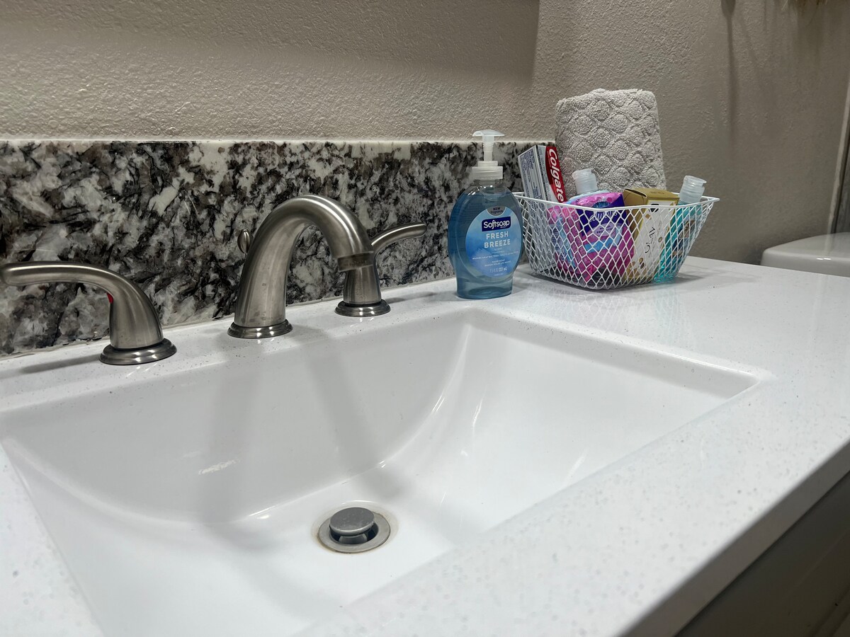 A white countertop features a sleek, undermount sink with polished chrome faucets. Nearby, a basket holds assorted toiletries and cleaning supplies. A roll of paper towels is neatly placed, complementing the modern, clean design of the bathroom space.