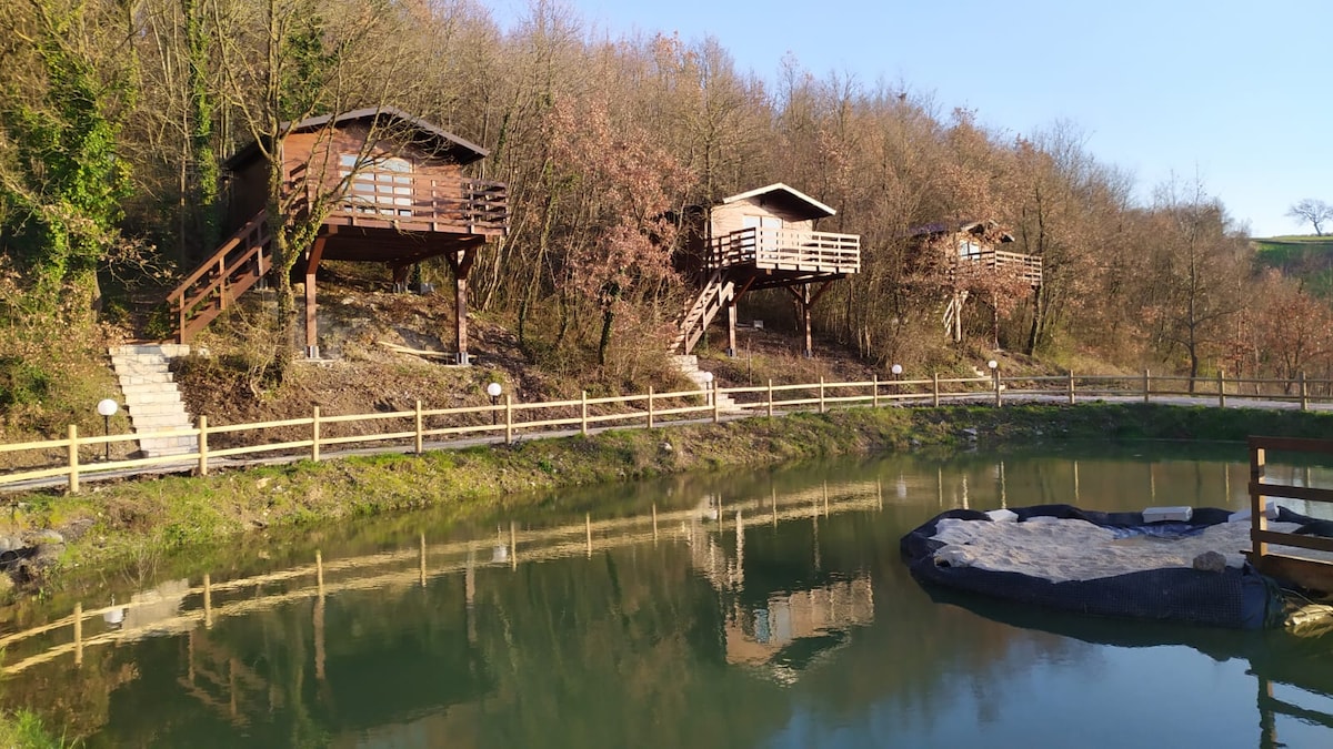 Three wooden tree houses are positioned on elevated platforms along the edge of a serene pond, surrounded by greenery. Wooden steps lead up to each tree house, which features balconies overlooking the water. Natural light reflects off the calm pond surface.