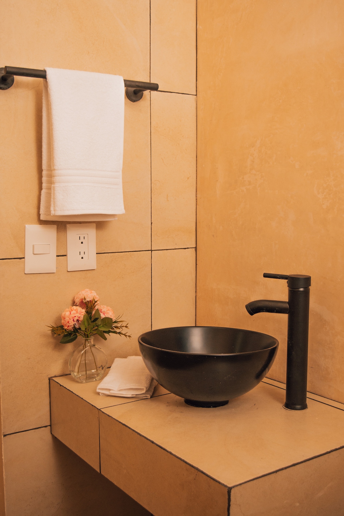 A minimalist bathroom features warm-toned walls, with a sleek black bowl sink and a contemporary faucet. A white towel hangs on a wall-mounted rack beside a small vase of pink flowers. A stack of white towels is neatly arranged on the surface below.