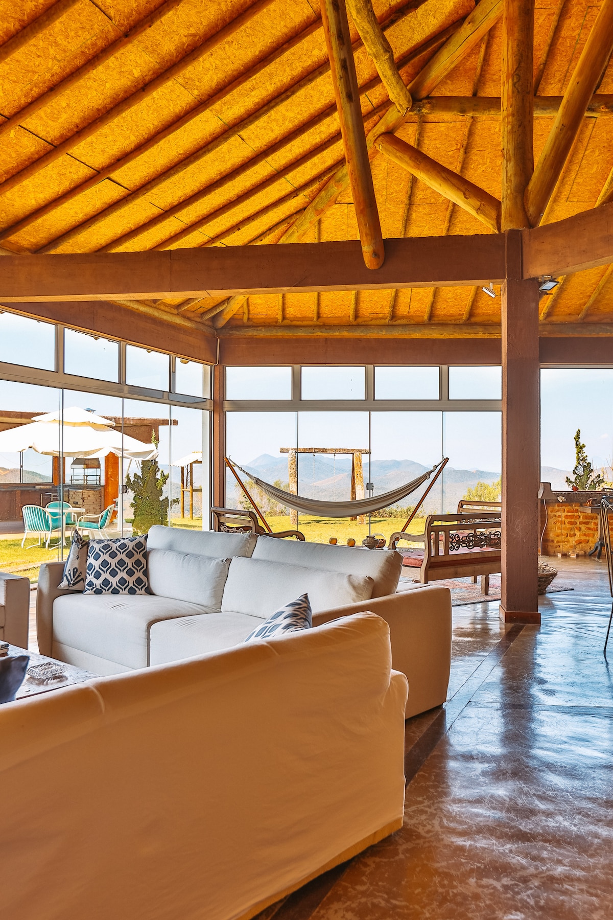 A spacious living area features a cozy white sofa under a natural straw ceiling, with large windows framing panoramic mountain views. A hammock is positioned for relaxation, while outdoor seating is visible through the glass doors, inviting fresh air and sunlight into the space.