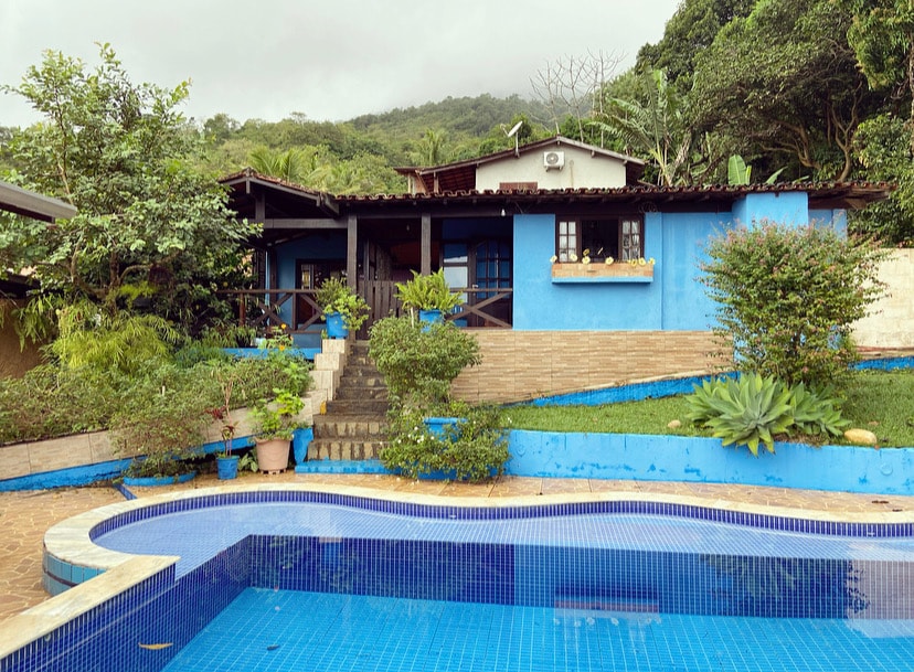 A charming house with blue accents is surrounded by lush greenery. The exterior features a spacious patio area, stone steps leading to the entrance, and a swimming pool with mosaic tiles. A sense of tranquility is emphasized by the natural landscaping.