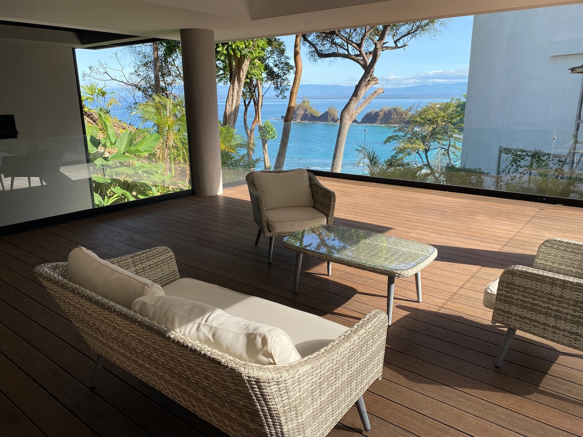 A spacious outdoor seating area features comfortable wicker chairs and a glass table, with a panoramic view of the ocean framed by lush greenery. The wooden deck creates a warm ambiance, providing an inviting space to relax while enjoying nature's beauty.