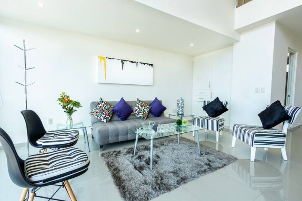 A modern living area features a gray sofa adorned with colorful cushions, complemented by two striped armchairs. A glass coffee table sits atop a soft area rug. Natural light filters through spacious windows, highlighting the bright decor and minimalist design.