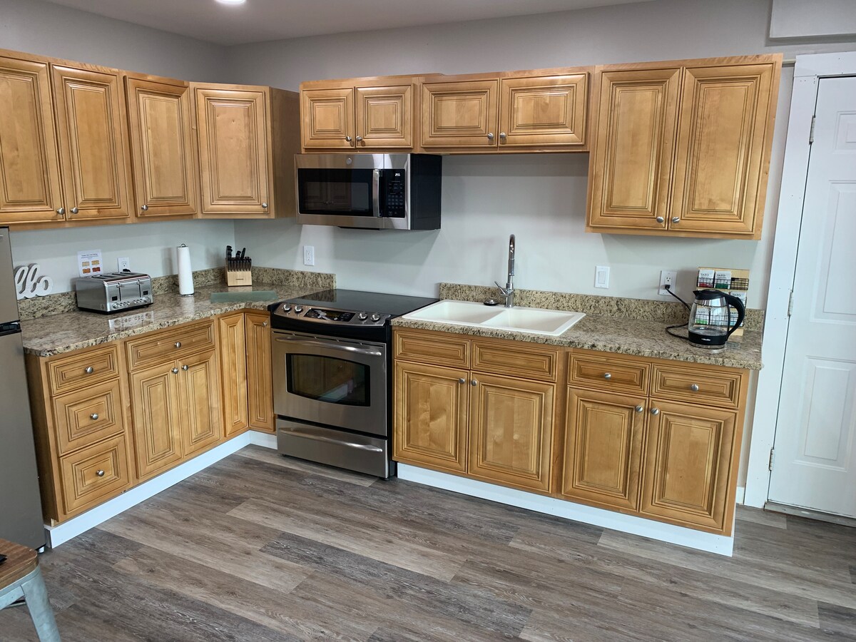 A modern kitchen setup features wooden cabinetry and granite countertops. Stainless steel appliances, including a microwave and a stove with an oven, are visible. A spacious sink is located near the countertop, which is well-organized, and a door leads to another area.