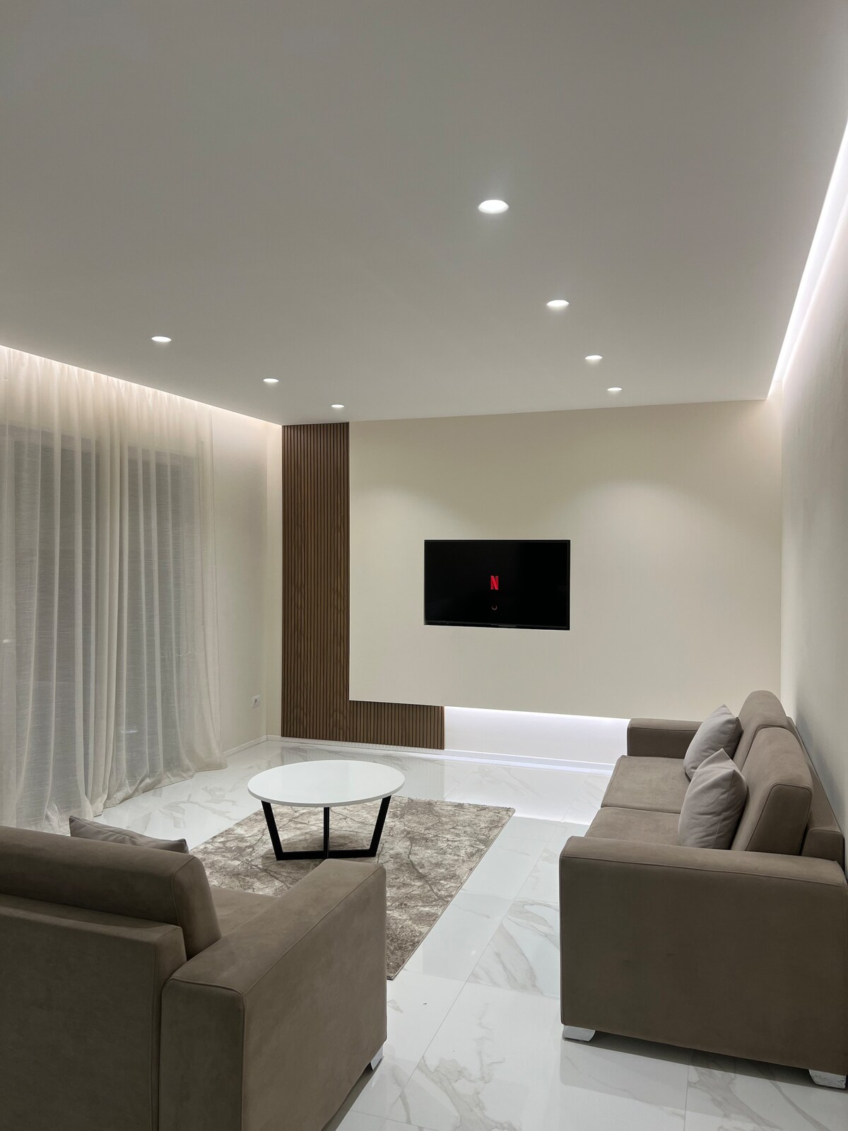 A modern living area features a light-colored sofa and armchairs arranged around a circular coffee table. A flat-screen TV is mounted on a simple wooden panel, and soft lighting is provided by recessed ceiling fixtures. Sheer curtains allow natural light to filter in.