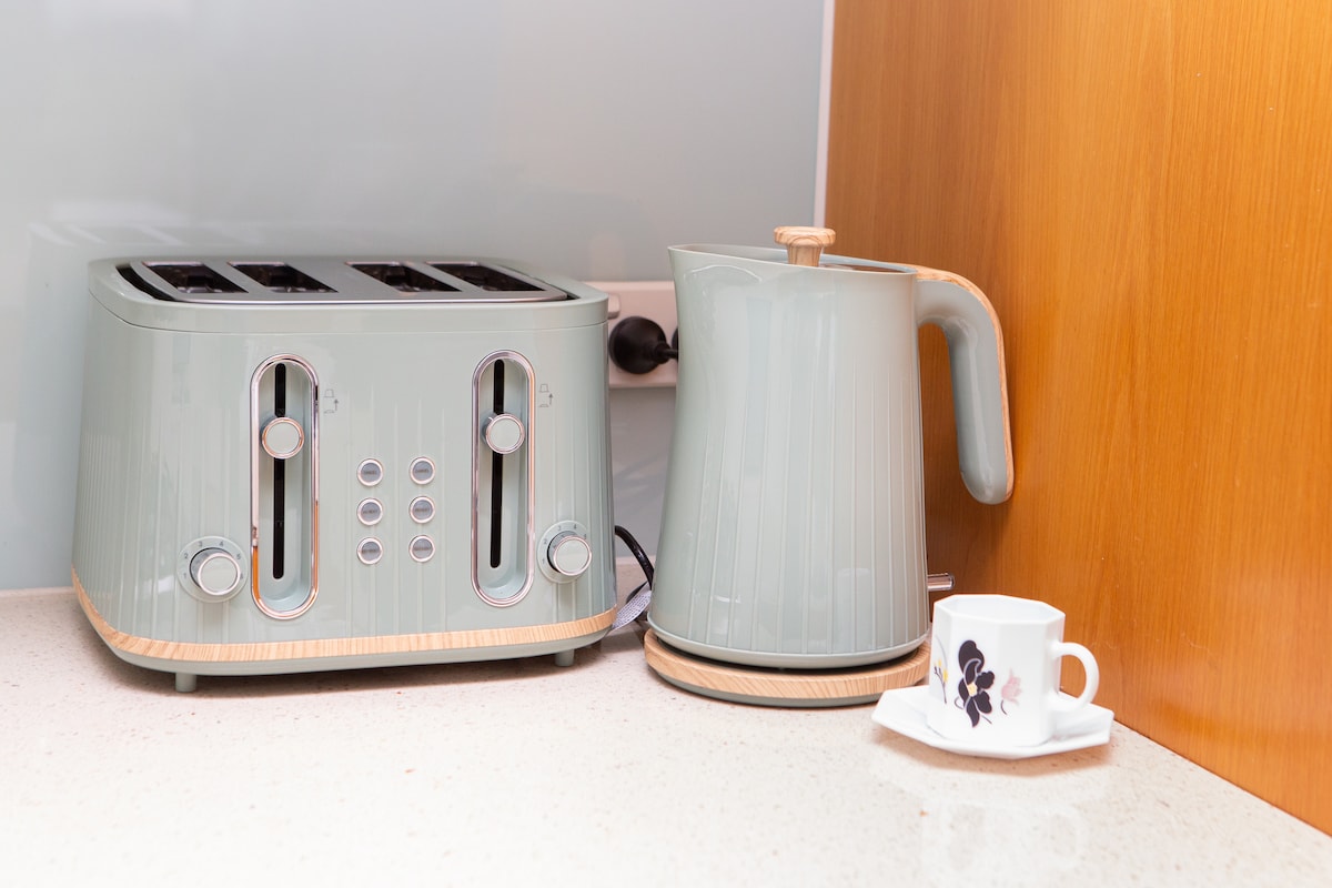 An elegant kitchen countertop displays a stylish green toaster and kettle, both featuring wooden accents. A white cup with a floral design rests nearby, adding a touch of charm to the modern kitchen setting.