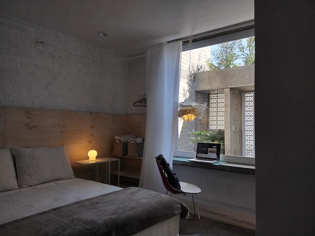 A modern bedroom features a spacious bed with neutral linens and a bedside lamp emitting soft light. A window offers natural light and views of greenery, while a small table with a laptop is positioned nearby, creating a functional workspace.