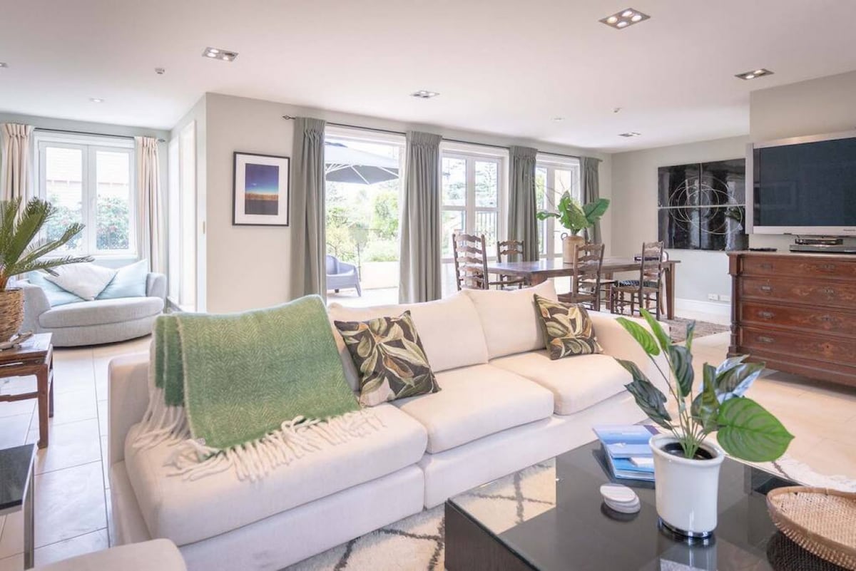 A spacious lounge is filled with natural light from large windows, creating an inviting atmosphere. Comfortable seating is complemented by a soft blanket and decorative cushions. A dining area is visible in the background, featuring a wooden table and chairs, with greenery subtly integrated into the decor.