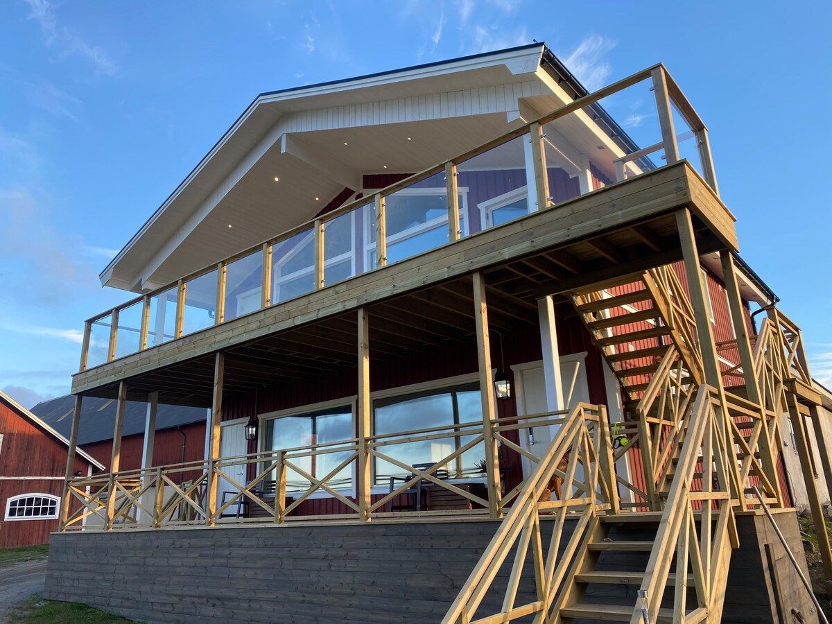 The exterior of a modern red house is displayed, featuring a large wooden deck with a spacious railing. Steps lead up to the entrance, and expansive glass windows illuminate the upper level, allowing for ample natural light.