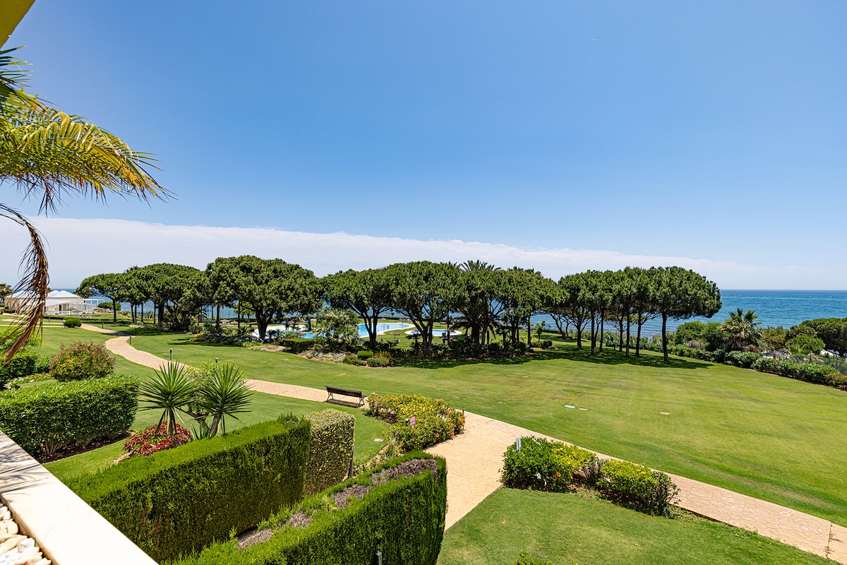 A wide view of meticulously landscaped gardens is presented, with vibrant greenery and palm trees lining the paths. The Mediterranean Sea is visible in the background, framed by a clear blue sky.