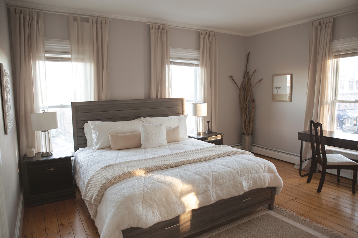 A spacious bedroom is presented with a king-size bed adorned with soft, white linens. Natural light filters through two windows, highlighting the warm wooden flooring. A wooden desk and chair are positioned against a wall, complemented by minimalist décor and a tall plant in the corner.