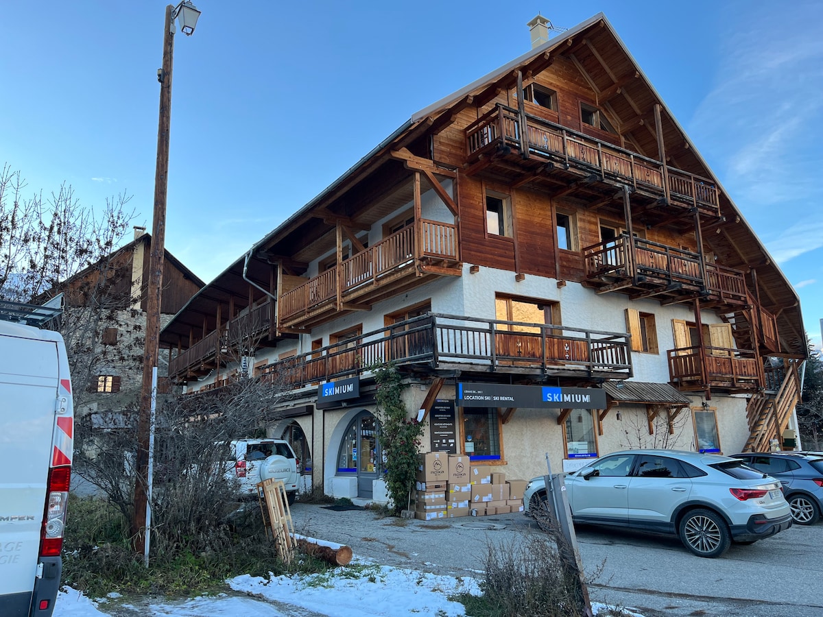 The exterior of a multi-level chalet features wooden balconies and snow-covered ground. Ground-level shops, including a ski rental shop, can be seen. A mix of vehicles is parked in front, with a mountainous backdrop under a clear blue sky.