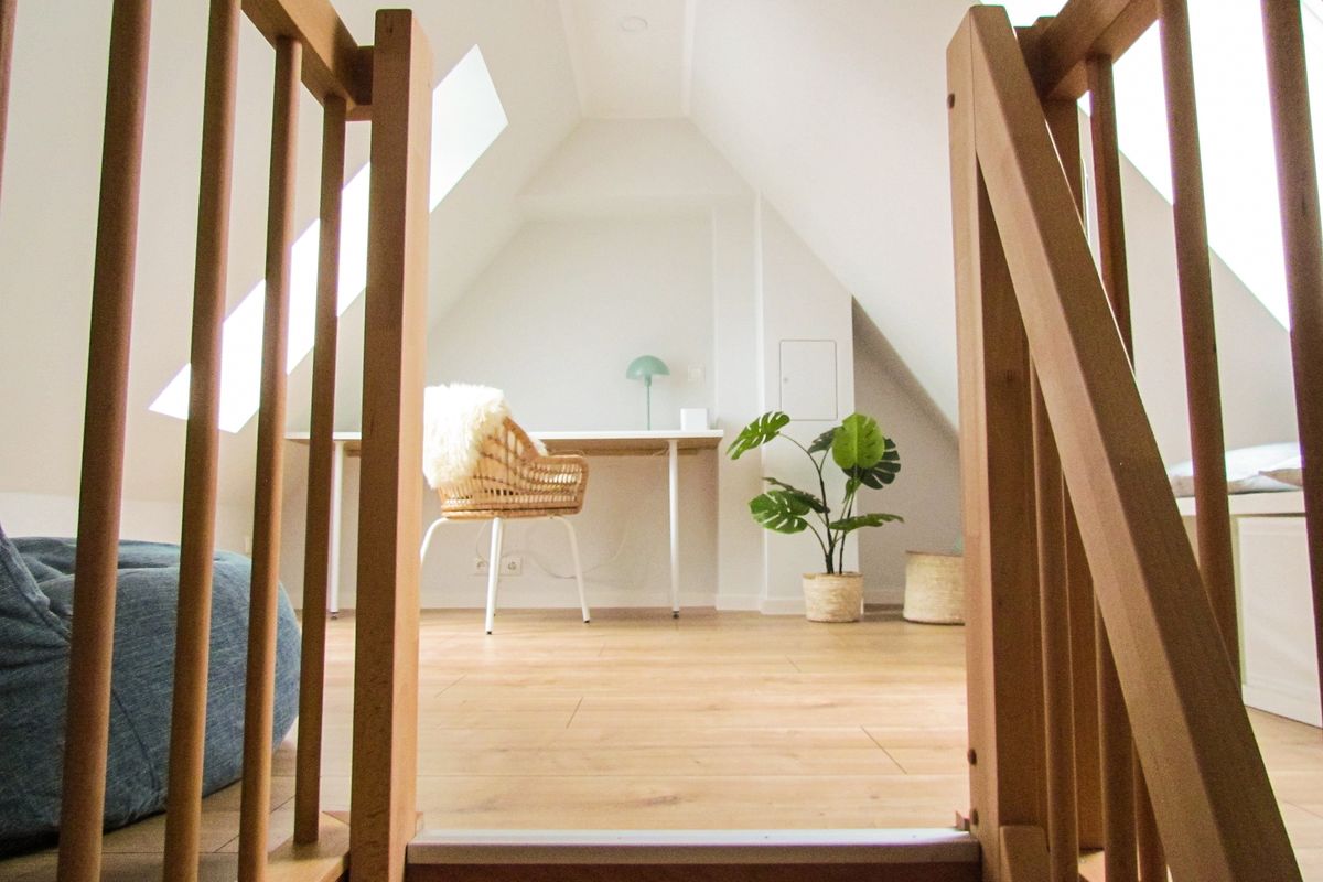 A bright loft area is seen, featuring a spacious wooden floor and sloped ceilings with two skylights. A minimalist desk with a chair is positioned against the wall, accompanied by a plant in a woven basket, creating an inviting workspace.