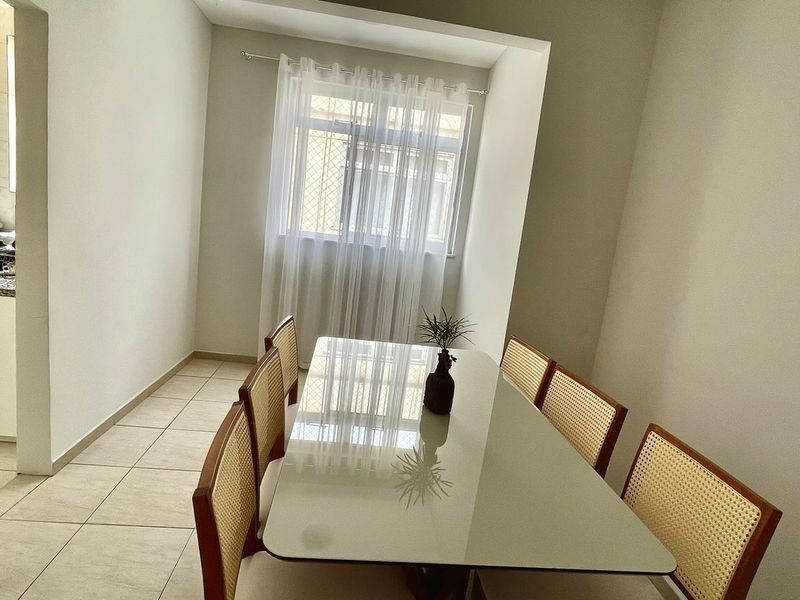 A dining area is presented with a rectangular table surrounded by six wooden chairs. A small potted plant occupies the center of the table. Natural light filters through sheer curtains on a window, creating a bright and airy atmosphere.