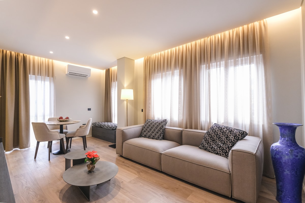 A modern living area is presented with a light-colored sofa and two accent chairs positioned around a circular coffee table. Natural light filters through sheer curtains draping over two windows. A decorative blue vase and a small floral arrangement add subtle color to the space.