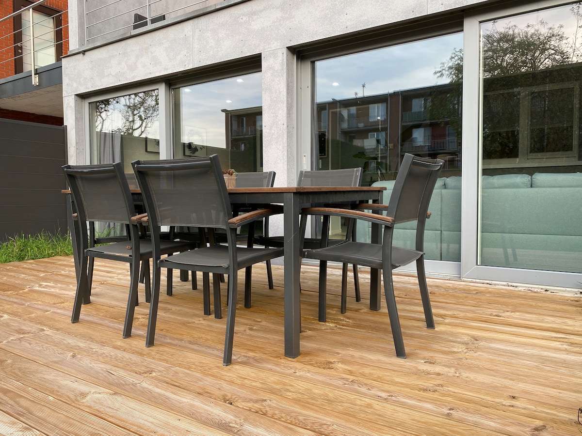 A wooden terrace features a dining table surrounded by six chairs, inviting outdoor meals. Large glass doors provide a view into the living space, enhancing the connection between indoor and outdoor areas.