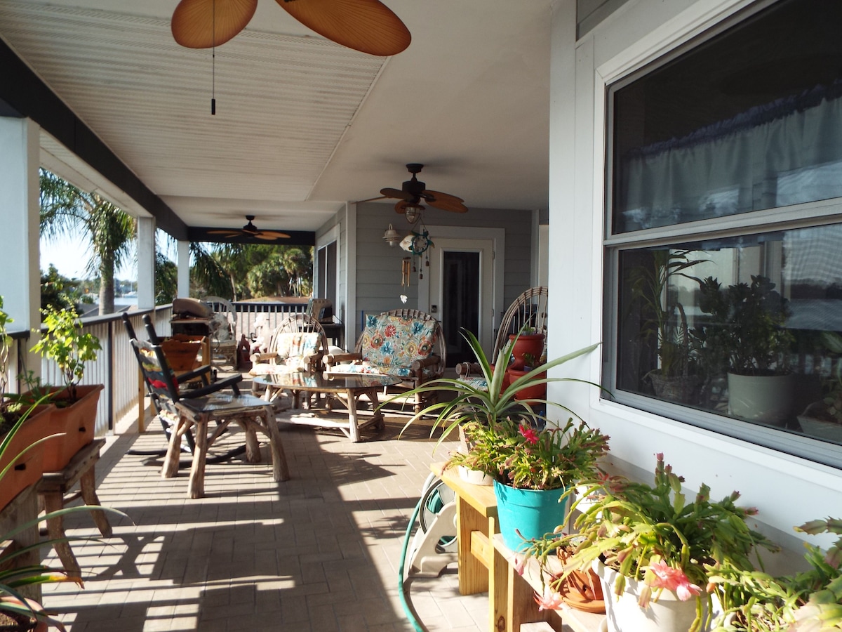 A spacious porch is adorned with various potted plants and comfortable seating arrangements, including chairs and a table. Ceiling fans provide airflow, while natural light filters through the windows, enhancing the inviting atmosphere. The view of the surrounding greenery adds to the serene setting.