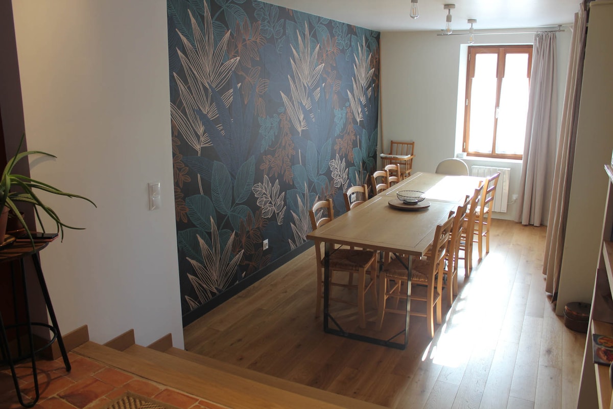 The dining area is highlighted by a large wooden table designed to seat ten. Natural light enters through a window, illuminating the space. A wall adorned with tropical foliage wallpaper adds a vibrant touch, while wooden chairs surround the table, creating an inviting setting.
