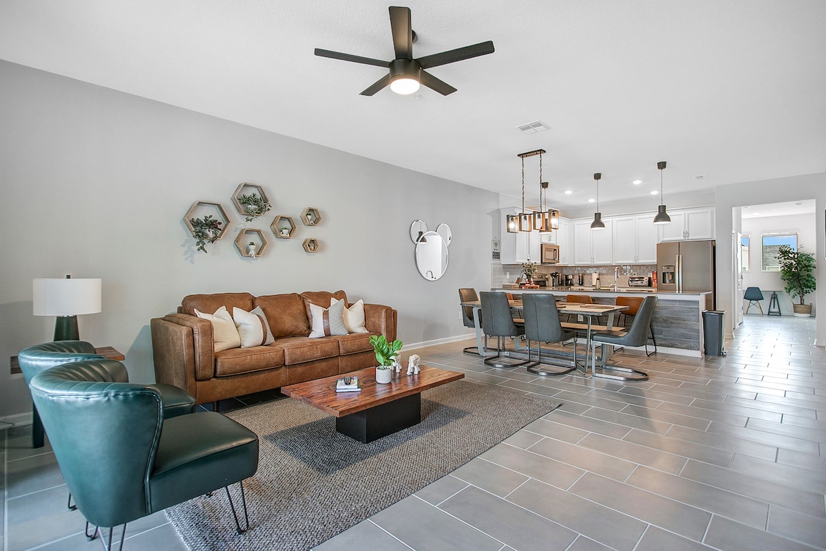 The living area features a brown leather sofa, two green accent chairs, and a wooden coffee table atop a grey rug. A dining area with metal chairs is visible in the background, along with kitchen cabinetry and pendant lighting that provides a modern touch.