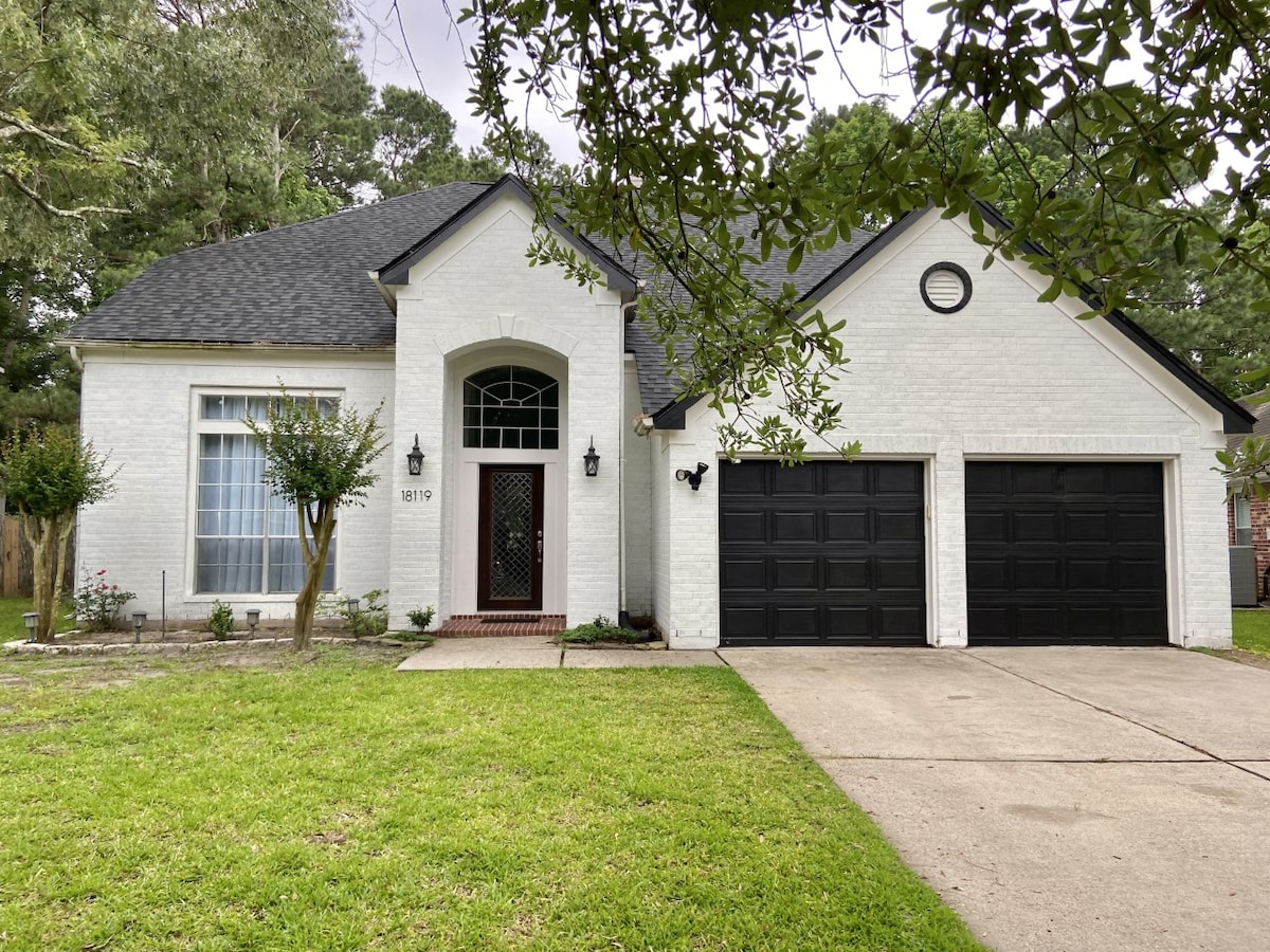 The exterior of a well-maintained white brick house is showcased, featuring a symmetrical facade and a welcoming entrance with a decorative door. Two black garages are visible, accompanied by a lush green lawn and landscaping that enhances the overall appeal.