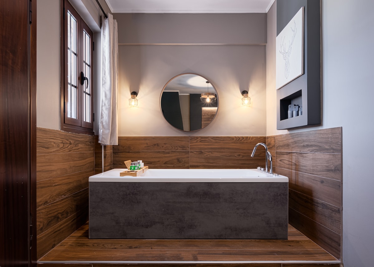 A well-appointed bathroom features a jacuzzi bathtub set against wooden paneling and grey walls. Natural light filters through a window, complementing the round mirror above the sink. Subtle lighting fixtures provide a warm glow to the inviting space.