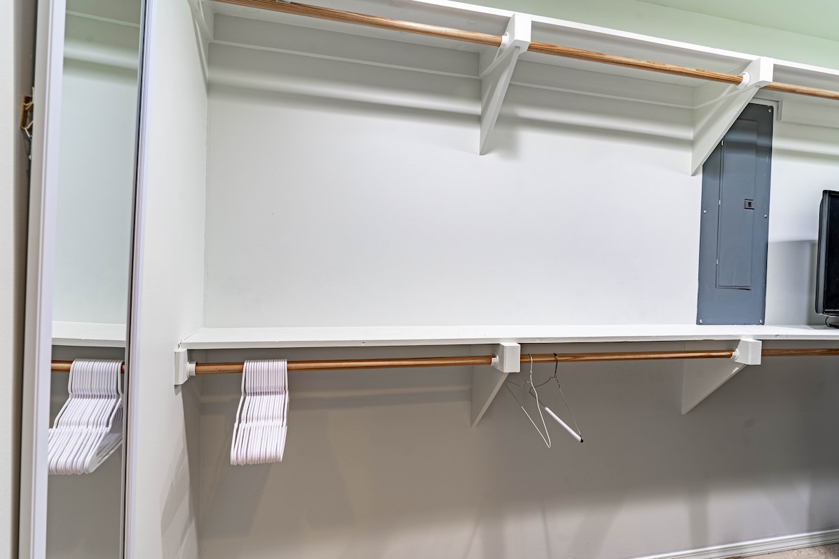 An uncluttered closet space is shown, featuring a long hanging rod supported by two shelves above. A few hangers are visible on the rod, while a small area on the right contains a power panel. The walls are painted in a neutral color.