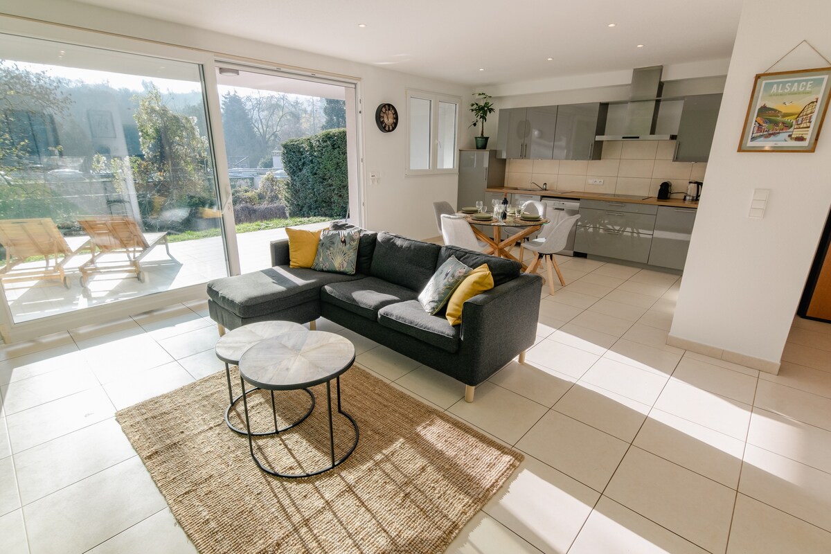 A modern living area features a gray sectional sofa complemented by vibrant cushions. A round coffee table sits on a natural fiber rug. Bright light enters through large sliding glass doors that open to a terrace with outdoor seating and views of the garden.