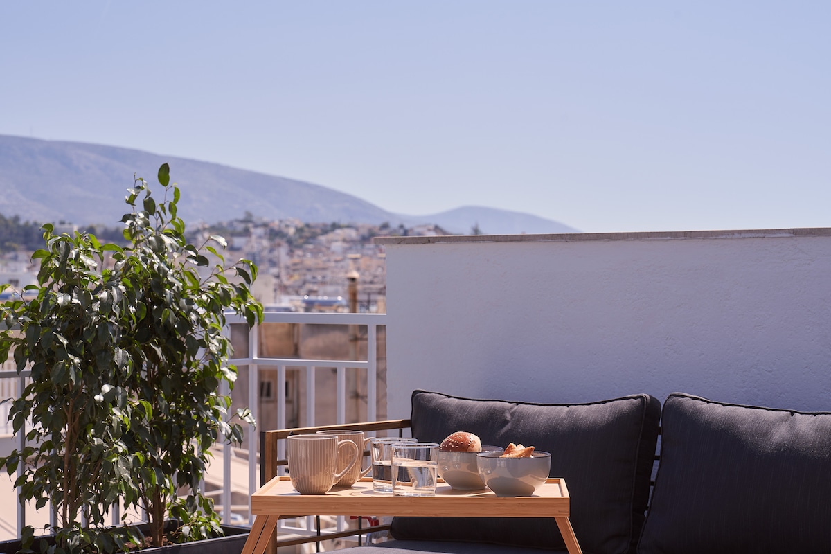 A sunlit terrace features a low wooden table, set with a pitcher of water, glasses, and bowls of food. A cozy seating area is adorned with dark cushions and surrounded by greenery, complemented by a scenic view of distant mountains and city rooftops.