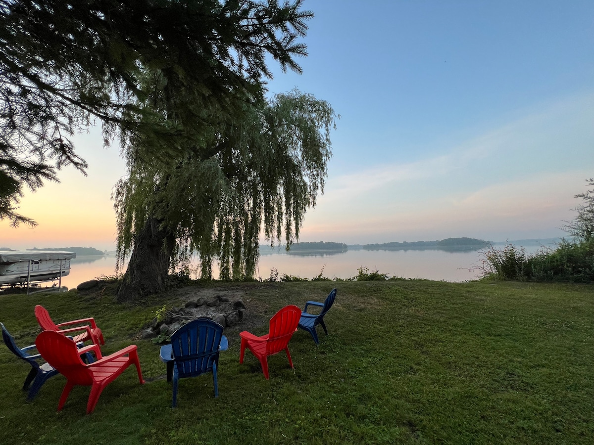 A peaceful outdoor setting is presented with colorful seating arranged around a fire pit, surrounded by lush grass. A large tree provides shade, and the calm water of Sinissippi Lake reflects the soft colors of dawn, offering a serene atmosphere for relaxation.